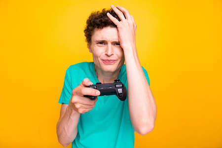 Handsome curly-haired upset young guy wearing casual green t-shirt, losing video game battle. Isolated over yellow backgroundの写真素材