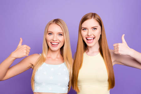 Advertisement concept. Portrait of positive pretty girls with long hair in casual outfits showing thumb up signs with hands looking at camera isolated on bright violet backgroundの写真素材