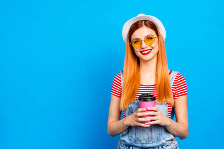Cute young girl looks into the camera and holds pink paper glass with a drink in her hands isolated...