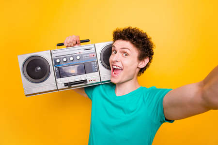 Young attractive handsome funky curly-haired excited guy wearing casual green polo t-shirt listening loud music, holding boombox. Isolated over bright shine vivid yellow backgroundの写真素材