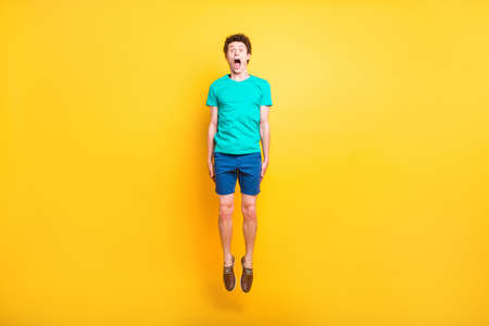 Full size length body picture of handsome curly-haired playful young guy wearing casual green t-shirt, shorts, shoes, jumping up in air, opened mouth. Isolated over yellow backgroundの写真素材