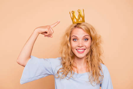 Portrait of young attractive charming nice cute caucasian curly-haired blonde girl wearing golden crown and blue dress, pointing to diadem, toothy smile. Isolated over beige backgroundの写真素材