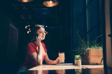 Attractive nice smiling young girl wearing casual sitting in cafeteria, talking on phone, drinking coffee, dark interiorの写真素材