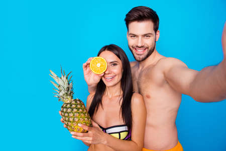 Handsome guy and brunet pretty woman make a selfie on phone over blue background. Summer portrait of hot, charming, tourists in swimsuits. Woman hold ananas on her hand, man keep orange on eye placeの写真素材