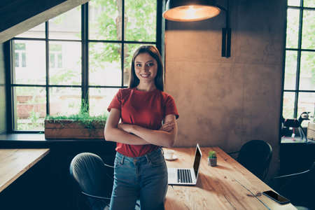 Cute and charming young woman in red t-shirt stands in cafe with arms crossed on chest, looks at camera and smilingの写真素材