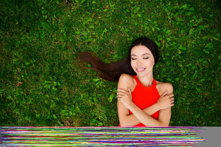 Caring for yourself is more important than any business! Asian girl in red dress, with dark hair lies on the grass with closed eyes in the park gently embracing herselfの写真素材