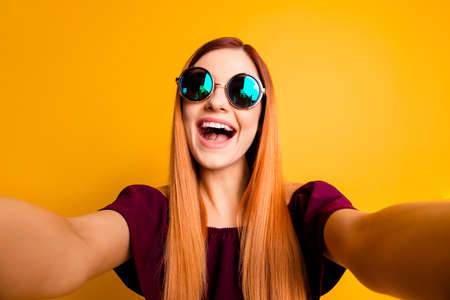 Close up portrait of silly mad girl in glasses with beaming smile making selfie on front camera of smartphone isolated on yellow backgroundの写真素材