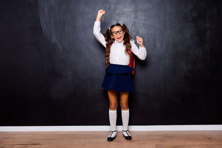 Full size body length of glad smart cute stylish small little girl with curly pigtails in white blouse shirt and blue skirt with red bag, raising hands up. Isolated over black backgroundの写真素材