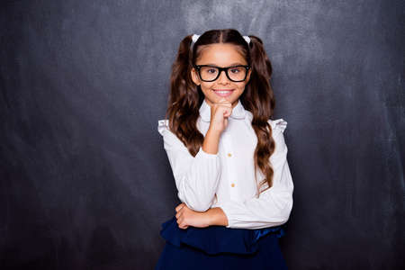 Portrait of positive glad nice adorable cute small little girl with curly ponytails in white formal blouse shirt, skirt, touching chin. Isolated over black backgroundの写真素材