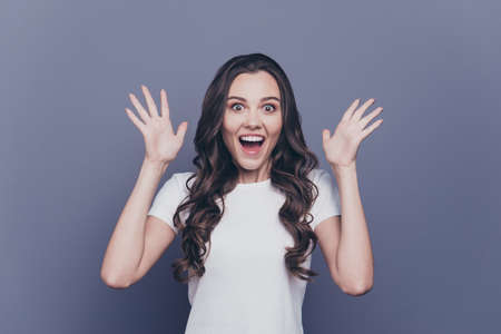 Attractive pretty charming adorable cheerful stylish brunette curly-haired girl in white t-shirt, showing wow facial expression and hands gesture, opened mouth, isolated over grey backgroundの写真素材