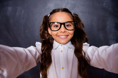Self-portrait of cheerful nice genius adorable cute lovely small little girl with curly ponytails in white formal blouse shirt, smiling to cam. Isolated over black backgroundの写真素材