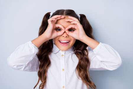 Portrait of nice cute funky cheerful adorable lovely stylish playful small, little girl with curly ponytails in white formal blouse shirt, showing ok-signs like glasses. Isolated over grey backgroundの写真素材