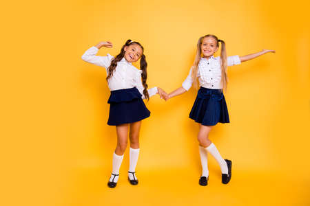 Back to school concept. Full length, legs, body, size portrait of  small girls happily jump holding hands isolated on bright yellow backgroundの写真素材