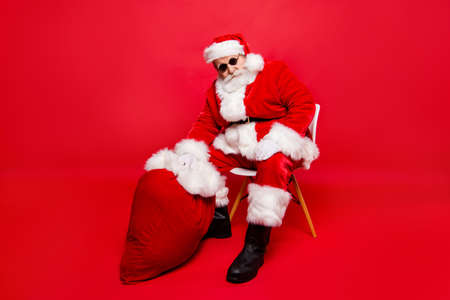 Winter Christmastime party over. Full legs body size confident calm serious Santa in shoes white gloves spectacles with beard sit on chair hold heavy sack by hand isolated on red shine backgroundの写真素材