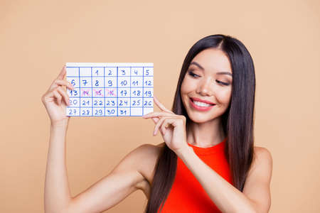 Close up photo portrait of pretty cheerful cute dreamy brunette brown haired lady holding showing handmade calendar in hand with three crossed days isolated on pastel beige backgroundの写真素材