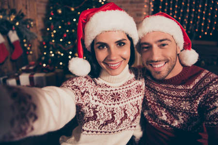 Winter christmastime concept. Handsome man and brunette beautiful, sweet, charming, attractive lady in ornament sweater stand in comfort, cozy house interior make selfie on front camera of smartphoneの写真素材