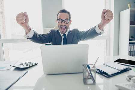 Confident director economist employee professional realtor man in formal wear sit on chair armchair behind table in white office look at modern technology monitor screen raised fists upの写真素材