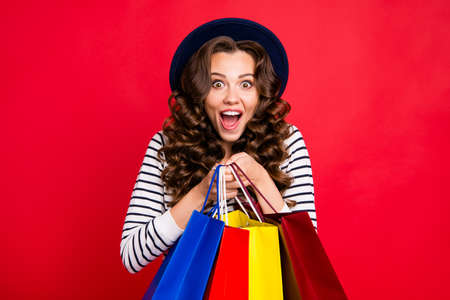 Portrait of nice glad amazed attractive pretty cheerful positive wavy-haired lady wearing striped pullover carrying colorful bags clothes accessory isolated over bright vivid shine red backgroundの写真素材
