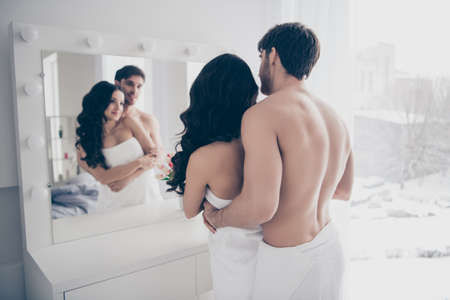 A couple in white towels hugging and looking at the mirror in the bathroom. back viewの写真素材