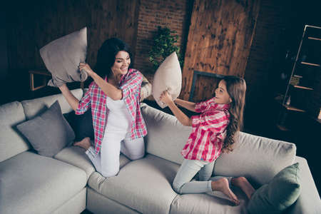 Close up photo cheerful two people mum little daughter yelling loud playful mood chill out pillow fight barefoot wear pink plaid shirts flat apartment room sit lying cozy couch sofa divanの写真素材