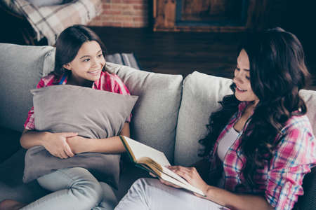 Close up photo of two people mum little daughter look open book hold hands arms adventure novel interesting listening wear pink plaid shirts flat apartment room comfortable cozy couch sofa divanの写真素材