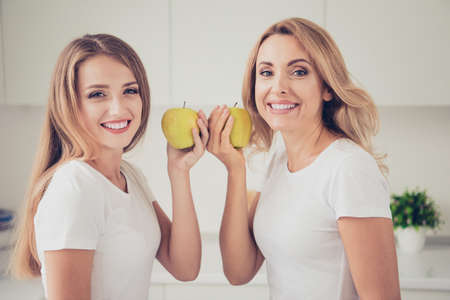 Close-up portrait of two nice-looking cute lovely attractive charming cheerful cheery blonde ladies holding in hands fresh natural apples in modern light white interior roomの写真素材