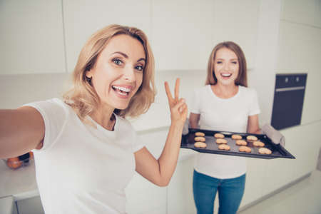 Close up photo showing v-sign two people mum and teen daughter enjoy spending time make take selfies hot heart shape figure cookies housewife help wear white t-shirts jeans in bright flat kitchenの写真素材