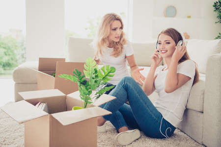 Close up photo two people mom teen daughter big boxes earflaps on head not listen mum lecture asking help wear white t-shirts jeans in bright flat sit in front comfortable sofa floor carpetの写真素材