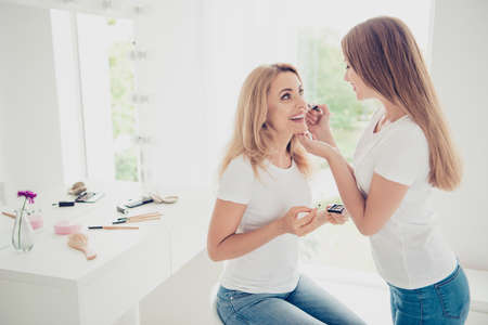Close up side profile view photo two people mum teen daughter excited glad weekend communicate doing indoors makeup applying cheekbone wear white t-shirts jeans bright flat apartment room placeの写真素材