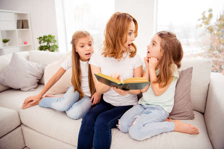 Portrait of nice cute amazed shocked lovely attractive charming cheerful cheery redhair people mom mommy mum reading book pre-teen girls sitting on divan in house indoorsの写真素材