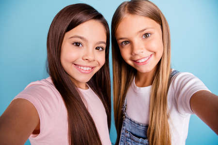 Close up photo two little she her blond brunette girls long pretty hair telephone make take selfies mom mommy skype talk tell speak wearing casual jeans denim t-shirts isolated blue bright backgroundの写真素材