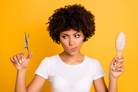 Close up photo beautiful amazing she her dark skin lady hold hair brush scissors hands pros cons ready change herself style wear casual white t-shirt isolated yellow bright vibrant vivid backgroundの写真素材