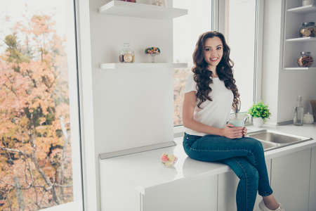 Close up photo beautiful curly brunette she her lady homey indoors hold hot beverage glad clean perfect white flat sit table wearing domestic home apparel clothes outfit comfy kitchenの写真素材
