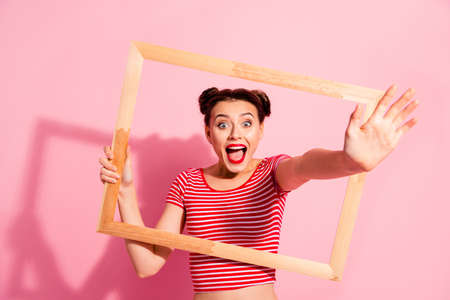 Portrait of her she nice cute charming attractive funny funky cheerful cheery girl in striped t-shirt holding in hands wooden frame opened mouth giving hand isolated on pink pastel backgroundの写真素材