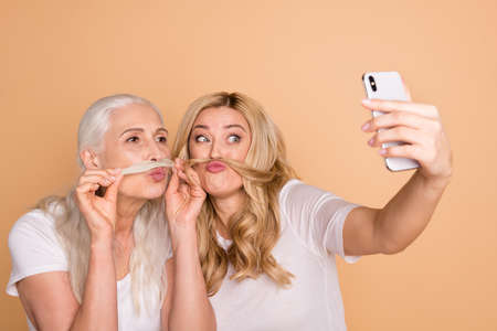 Close-up portrait of nice lovely attractive charming cute cheerful cheery playful comic foolish crazy ladies wearing white t-shirt taking making selfie curls isolated on beige pastel backgroundの写真素材