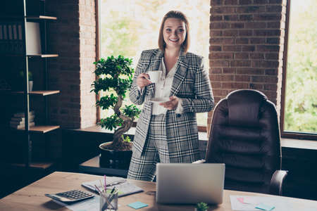 Close up photo beautiful amazing she her business lady hold white glass cup hot beverage cheerful look guests invite come in notebook table sit office wearing specs formalwear checkered plaid suitの写真素材