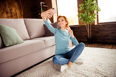 Close up photo beautiful she her lady hand arm telephone make take selfies show v-sign say hi wear blue pullover jeans denim clothes sit floor fluffy carpet divan sofa couch house living room indoorsの写真素材