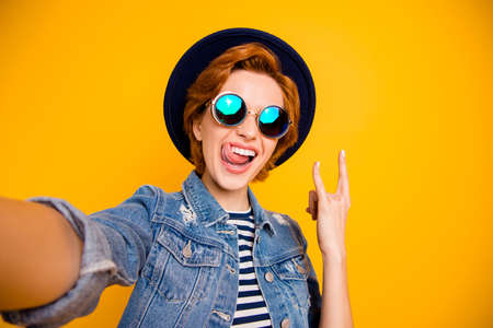 Close up photo funny funky foxy she her lady make take selfies metal music lover tongue out mouth wear specs vintage hat casual striped t-shirt jacket jeans denim isolated yellow bright backgroundの写真素材