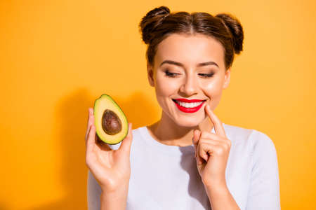 Close up photo beautiful she her lady pretty hairdo hold slice avocado look side with interest hand arm finger chin yummy tasty wear casual white pullover clothes isolated yellow backgroundの写真素材
