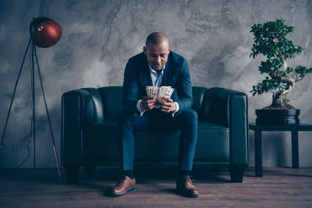 Portrait of nice classy elegant attractive busy guy wearing jacket suit top manager sitting on couch account budget balance at workplace station gray concrete wall.の写真素材