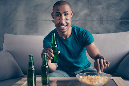 Close up photo amazing he him his dark skin macho handsome hold arms hands green bottle ale cider potato fried fast food pause break wear blue t-shirt pants sit comfy divan room office house indoorsの写真素材