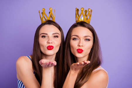 Close up portrait two people beautiful she her models chic ladies hands arms palms send air kisses servants gold crowns head wear summer colorful dresses isolated purple violet bright backgroundの写真素材