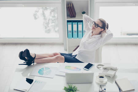 Close up photo beautiful she her business lady hands arms behind head joy fun overjoyed vacation thoughts telephone notebook papers legs heels table wear specs formal-wear shirt skirt bright officeの写真素材