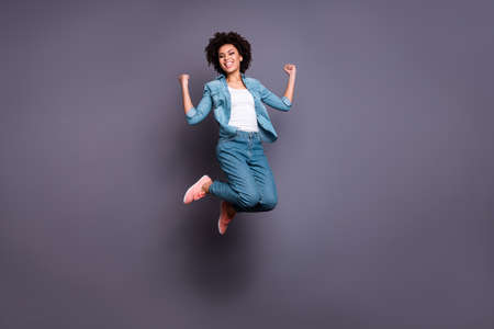 Full length body size photo beautiful she her dark skin lady jump high arms hands raised up air champion triumphant glory luck yeah wear casual jeans denim shirt isolated grey backgroundの写真素材