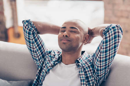 Close up photo of lovely nice man student hipster touch hands head palms close eyes sit divan sofa couch dressed checkered shirt big apartment interior quiet free timeの写真素材