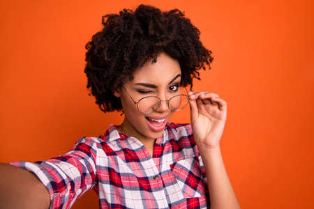 Photo of beautiful dark skin lady make selfies playful winking eye coquette wear specs casual checkered shirt isolated orange bright backgroundの写真素材