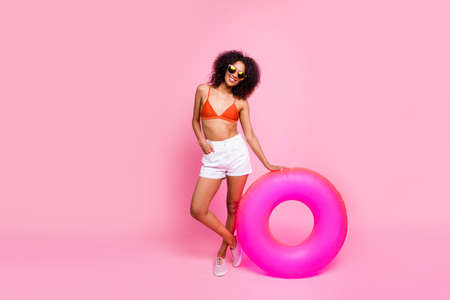 Full length body size photo beautiful she her dark skin lady enjoy seaside hold circle lifebuoy buoy ready try water wear sun specs swimming orange suit brassiere white shorts isolated pink backgroundの写真素材