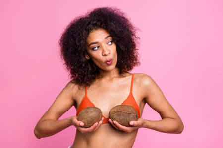 Close up photo beautiful funny she her dark skin bright lady stylish trendy hands arms hold coconuts try on new breast size chest look mirror wear swimming orange suit isolated pink backgroundの写真素材