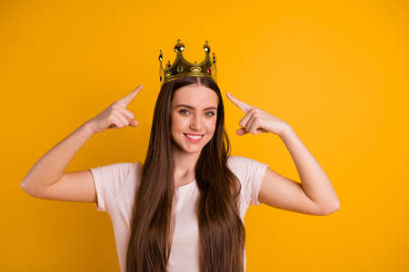 Close up photo of lady leader in gold crown on her head. Her fingers indicate special status of  pop star. Girls run the world concept. Wearing casual pastel t-shirt isolated yellow bright backgroundの写真素材