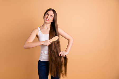 Close up photo beautiful mad  lady hold arms hands very long brown hair bad condition mat elf-lock curls hair brush angry wear casual white tank-top jeans denim isolated pastel beige backgroundの写真素材